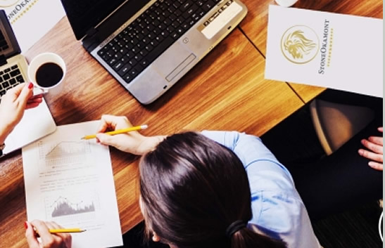 Imagem de uma mesa com notebooks, folhas de papel e uma pessoa olhando para um papel com gráficos